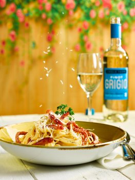 Food photograph of a bowl of spaghetti carbonara, with crisp bacon garnished with parsley and parmesan. Served on a white tabletop with a bottle and glass of white wine, in an outdoor setting with a fence and flowers in the background