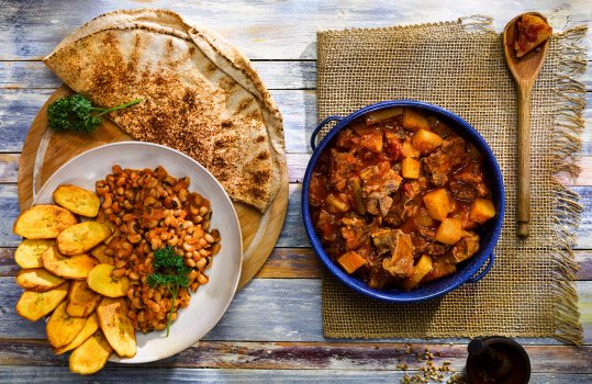 Aerial food photograph of Nigerian spicy black eye beans served with fried plantain and roti in an off white bowl on a wooden board, alongside turnip and lamb stew served in a deep blue enamel dish on top of a piece of hessian, all shot on a blue painted background