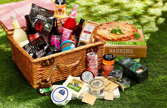 Food photograph of a wicker picnic hamper full of artisan Welsh food, including cheeses, beef jerky, honey & beer, among other things, shot in an outdoor setting on grass