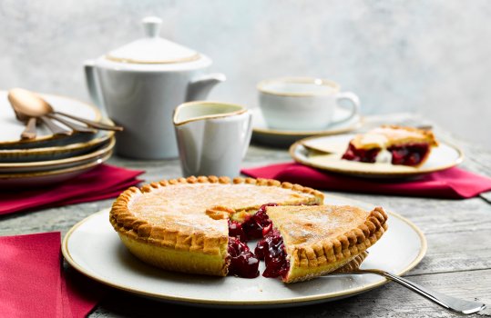Food photograph of a whole cherry pie shot on a white plate with a slice being removed to show the glossy cherry filling, served alongside a teapot and cups of tea