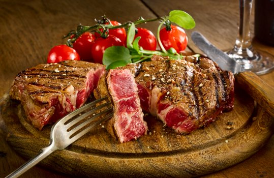 Food photograph of a juicy rare beef rib eye steak, with a crunchy crisp bar marked experior and a juicy pink inside. Served on a wooden board with watercress and vine cherry tomatoes, on a wooden tabletop