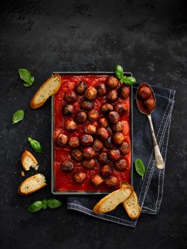 Aerial food photograph of a tray of meat free meatballs in a tray of rich tomato sauce, alongside toasted slices of ciabatta and sprigs of basil, shot on a black slate background