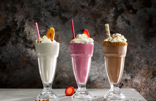 Food photograph of three milkshakes, banana, strawberry & chocolate milkshakes in sundae glasses shot on a dark grey background