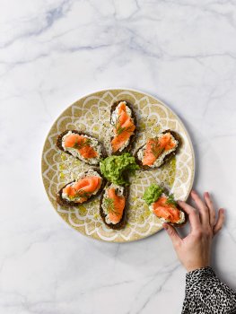 Aerial food photograph of smoked salmon and crushed avocado on top of dark Nordic style bread, arranged on a plate against a marble background, with one slice being removed by a woman’s hand