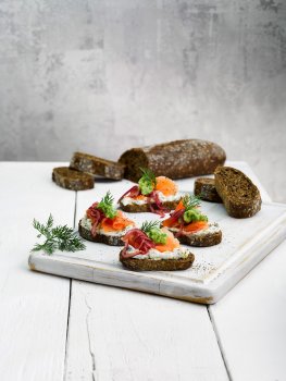Food photograph of smoked salmon and crushed avocado on top of dark Nordic style bread, arranged on a white chopping board with the rest of the loaf of bread, shot against a white and grey background