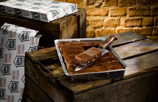 Food photograph of a chocolate topped peanut chocolate brownie, a traybake in a foil tray with a slice removed and presented on a chefs knife, served on a vintage apple crate with retail packaging in the background