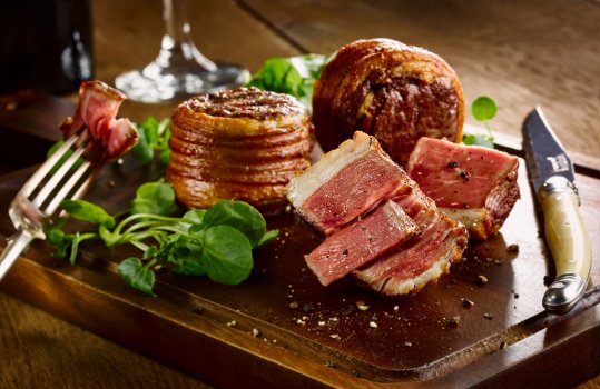 Food photograph of juicy rare lamb noisettes, with a crunchy crisp bar marked experior and a juicy pink inside. Served on a wooden board with watercress and vine cherry tomatoes, on a wooden tabletop