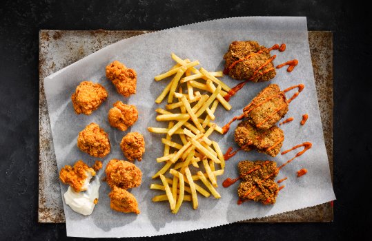 Aerial food photograph of a selection of pieces of southern fried chicken alongside fries, shot on a sheet of baking paper on a black slate background
