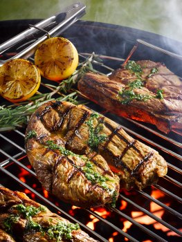 Food photograph close up of barbecued bar marked lamb leg steaks, topped with pesto sauce and shot alongside grilled lemons, on the grill of a lit barbecue in an outdoor setting