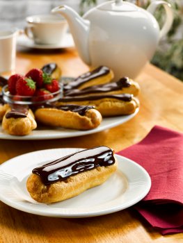 Food photograph of a shiny chocolate eclair on a white plate with a platter of eclairs garnished with strawberries in the background, along with a white teapot and cup and saucer, on a wooden tabletop with a white wall and a plant in the background