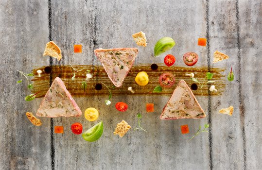Aerial food photograph close up of triangular slices of pancetta wrapped ham terrine shot on a glass background with scattered heritage baby tomatoes and micro herbs
