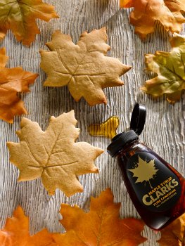 Aerial food photograph close up of two maple biscuits in the shape of maple leaves, shot on a light wooden background alongside a bottle of maple syrup, and real maple leaves
