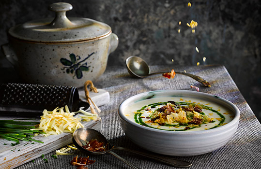 Food photograph close up of a bowl of smooth smoked cheese and potato soup, topped with crispy bacon, cheese crisps and vibrant green chive oil, shot in a dark atmospheric setting