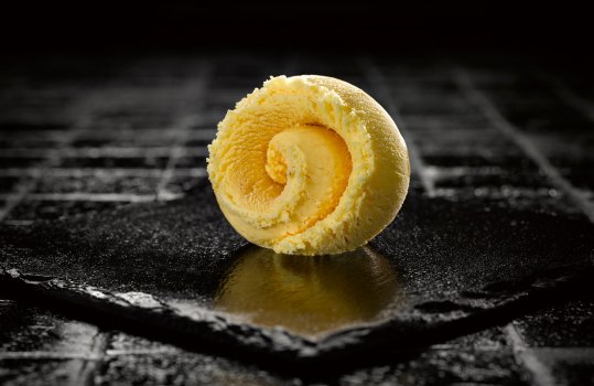 Food photograph close up of a curled scoop of tiramisu flavour gelato, shot on a sheet of slate against a shiny black background