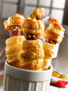 Food photograph close up of a Halloween party food recipe, hot dogs wrapped in a spiral of golden puff pastry with dots of mustard for eyes, served in a white ramekin in a home style setting