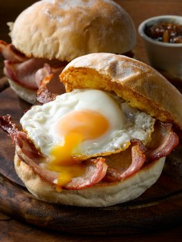 Food photograph close up of a toasted crispy bacon and fried egg sandwich, with the yolk broken and running out of the sandwich onto the dark wooden serving board