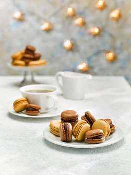 Food photograph close up of a plate of festive macarons. Chocolate, caramel and coffee flavoured macarons stacked on a white plate alongside a cup of coffee, shot on a white stone set with Christmas lights
