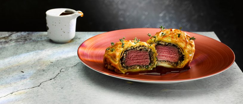 Food photograph close up of a fine dining main course, an individual beef wellington with golden flaky pastry and medium rare beef within, studded with thyme leaves. Served on a metallic orange plate alongside a small jug of red wine sauce