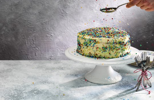 Food photograph of a vegan birthday cake, served on a cake stand and covered on all sides with sprinkles. Sprinkles are being spooned over the top and bouncing all over the image