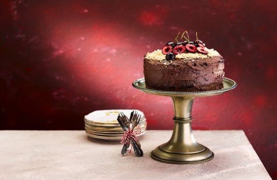Food photograph of a vegan birthday cake, served on a cake stand and covered with chocolate buttercream, topped with halved cherries and shot on a dark red setting