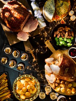Aerial food photograph of a Christmas dinner spread including Christmas ham, roast turkey, roast potatoes, roast parsnips, pigs in blankets, Brussels sprouts, Stilton cheese and a festive chocolate dessert. Shot on a dark background with fairy lights and Christmas crackers
