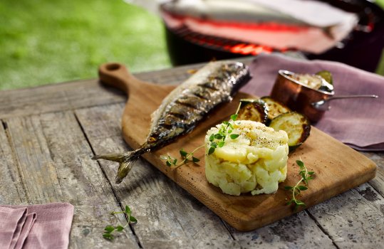 Food photograph of a barbecued mackerel served with crushed Pembrokeshire Early potatoes, shot in an outdoor setting with a lit barbecue with another mackerel in the background