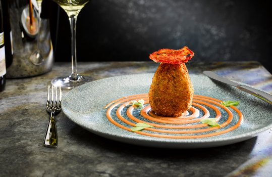 Food photograph of a fine dining starter, a Monmouthshire pork scotch egg served with a swirl of bloody Mary sauce, and garnished with celery leaves. Shot on a dark stone background with a tall glass of white wine
