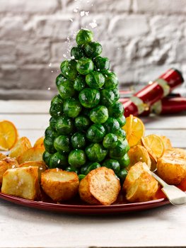 Food photograph of a Brussels sprout Christmas tree, bright green steamed sprouts arranged into a pyramid and surrounded by golden roast potatoes and Christmas crackers, shot in a white home style setting