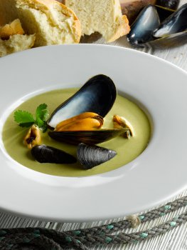 Food photograph close up of a restaurant dish, a green broccoli soup with parsley sprigs and mussels, crowned with a large mussel in its shell in the middle, shot with bread and scattered mussel shells in the background