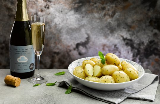 Food photograph close up of a bowl of buttered Pembrokeshire Early potatoes with herbs, shot in a dark setting alongside a bottle and glass of PGI Welsh wine