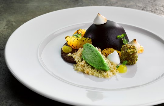 Food photograph of a fine dining dessert, a chocolate covered chocolate and orange mousse dome with charred orange supremes, pistachio stollen, basil sorbet and orange gel garnished with mini meringues and orange biscuit crumb. Served on a white plate on a concrete tabletop
