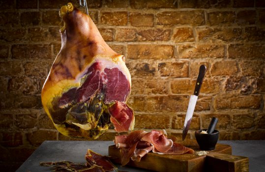 Food photograph of a whole leg of cured Carmarthen Ham, hanging from a butchers hook on a chain, with slices on a dark vintage wooden board