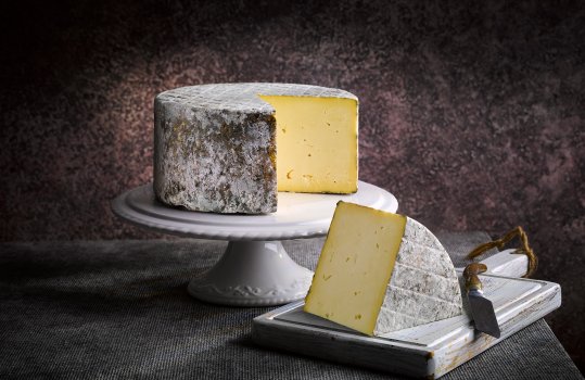 Food photograph of a whole Welsh PGI traditional Caerphilly cheese, the whole wheel is presented on a white ceramic cake stand with a wedge cut out and shown in the foreground on a white wooden board, shot against a dark background