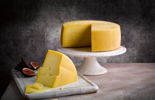 Food photograph of a whole Welsh PGI traditional Caerphilly cheese, the whole wheel is presented on a white ceramic cake stand with a wedge cut out and shown in the foreground on a white wooden board, shot against a dark background