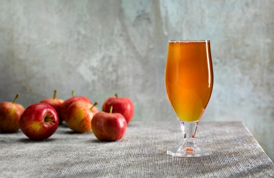 Drinks photograph of a glass of PGI traditional Welsh cider, shot on a textured grey and teal background with fresh bright red apples in the background