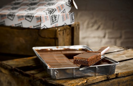 Food photograph of a chocolate and caramel topped fudgy chocolate brownie, a traybake in a foil tray with a slice removed and presented on a chefs knife, served on a vintage apple crate with retail packaging in the background