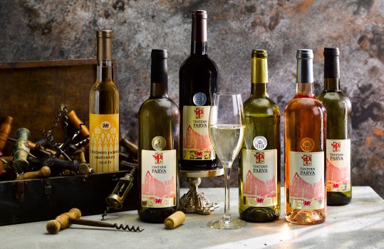 Drinks photograph of a group of bottles and a tall glass of PGI Welsh wine, shot on a concrete background against a mottled grey wall, alongside a box of vintage corkscrews