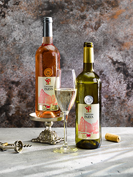 Drinks photograph of two bottles and a tall glass of PGI Welsh wine, shot on a concrete background against a mottled grey wall, alongside a vintage corkscrew