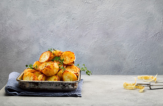 Food photograph of a heap of crispy golden roasted Welsh potatoes, shot on a grey background in a metal tray, alongside potato peelings and sprigs of thyme