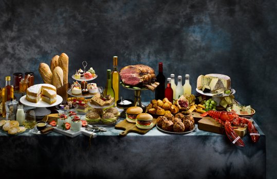 Food photograph of a groaning banquet table with a wide selection of Welsh produce, including cakes and patisserie, cheese, lobster, meat and poultry, pies, baked goods, world foods, wine and soft drinks