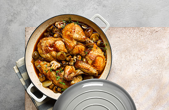 Aerial food photograph of a light grey casserole dish of chicken chasseur, bone in caramelised chicken pieces in a rich glossy sauce with mushrooms and baby onions, shot on a pink grey background