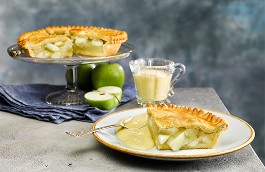 Food photograph close up of a slice of apple pie with a short golden pastry lid and a thick layer of apple filling, served on a plate with custard, with fresh green apples, and the rest of the apple pie in the background