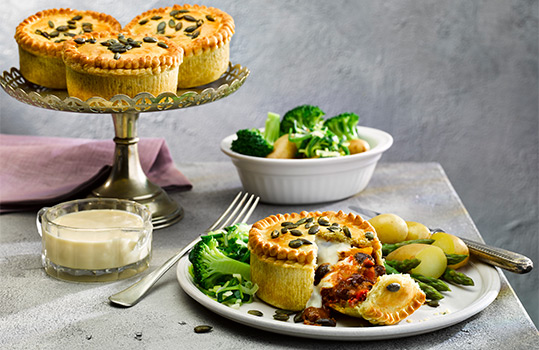 Food photograph close up of an individual butternut squash and cheese pie, with a short golden pastry lid topped with crispy pumpkin seeds, served on a plate with potatoes and green vegetables, with three more pies on a cake stand in the background