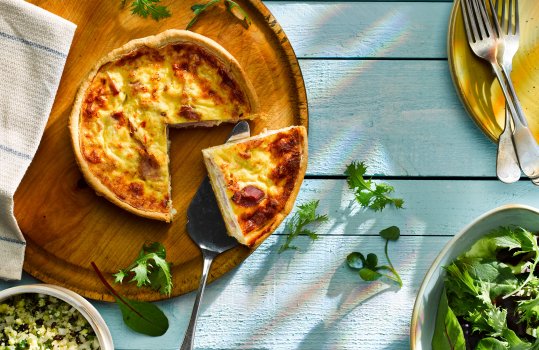 Aerial food photograph of a bacon and cheddar quiche with a slice being removed, shot on a wooden background with a pile of plates and forks, alongside bowls of salad