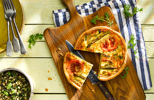 Aerial food photograph of a asparagus and tomato quiche with a slice being removed, shot on a wooden background with a pile of plates and forks, alongside bowls of salad