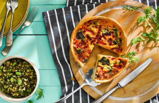 Aerial food photograph of a spinach and goats cheese quiche with a slice being removed, shot on a wooden background with a pile of plates and forks, alongside bowls of salad