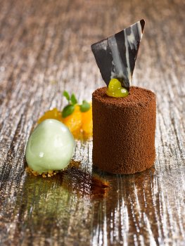 Food photograph of a fine dining dessert, a chocolate mousse cylinder coated in chocolate and chocolate powder, with orange gel, chocolate shard and a rocher of vanilla ice cream, served on a sheet of glass on a gold glitter tabletop