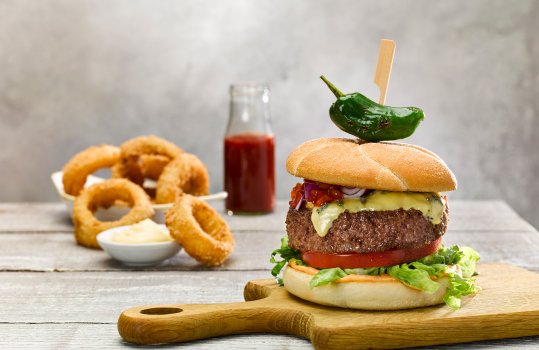 Food photograph close up of a thick grilled beef burger topped with melted blue cheese and red onion, served with fresh tomato and lettuce on a wooden chopping board, alongside panko breaded onion rings and grilled padron chilli peppers