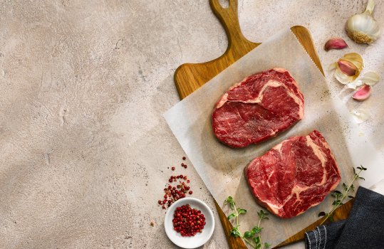 Aerial food photograph of two thick rib eye steaks, shot on a wooden chopping board with scattered pink peppercorns, thyme and garlic cloves