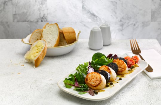 Food photograph close up of a plate of seared scallops and black pudding served with salad and shot on a grey stone background alongside a bowl of crusty bread